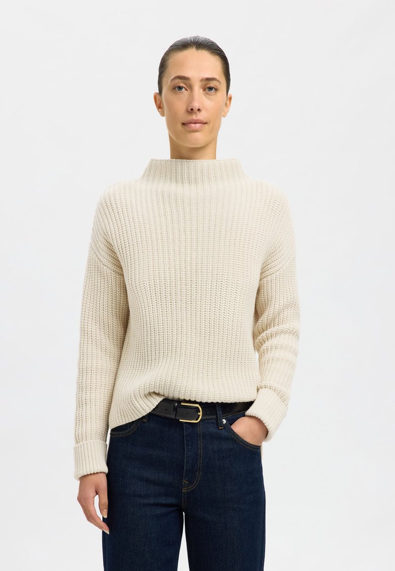 Helles beige, grob gestricktes Sweatshirt mit hohem Kragen, gerippter Textur und langen Ärmeln, kombiniert mit dunkelblauen Jeans und einem schwarzen Gürtel.