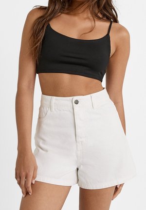 Femme portant un top court noir à fines bretelles et un short en denim blanc taille haute avec bouton à l'avant et poches.