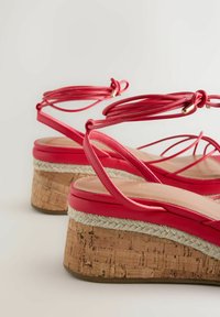 Sandales compensées rouges à semelles en liège, bordure en jute tressée, et brides autour de la cheville avec embouts dorés sur un fond clair.