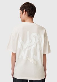 Camiseta de algodón de gran tamaño en color crema, con un gran texto gráfico blanco texturizado en la parte posterior. Mangas cortas y cuello redondo.