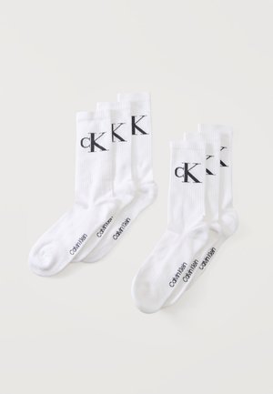 Dwa zestawy po trzy białe skarpetki Calvin Klein z czarnym logo "CK" na mankiecie i napisem "Calvin Klein" na stopie, ułożone na białej powierzchni.