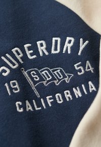 Granatowy materiał z białym haftowanym napisem "SUPERDRY 1954" i flagami; fakturowana powierzchnia; kontrastujący kremowy akcent na krawędzi.
