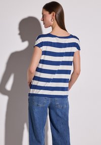 Top de manga corta y cuello redondo en rayas horizontales azules y blancas; tejido suave; detalle fruncido en los hombros; combinado con vaqueros azules.