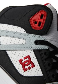 Black, white, and gray sneaker with perforated leather, red accents, and embroidered logo. Features a padded collar and lace-up design.