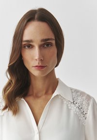 Camisa blanca de botones con bordado floral en el hombro. Presenta un cuello clásico y una tela suave y ligera con un corte relajado.