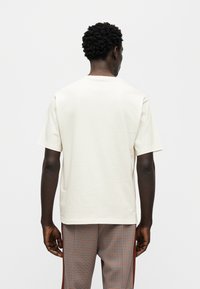 Κοντομάνικο, εκρού t-shirt με στρογγυλή λαιμόκοψη, κατασκευασμένο από βαμβάκι. Απλός σχεδιασμός, χωρίς ορατά μοτίβα ή διακοσμητικά, εμφανιζόμενο από πίσω.