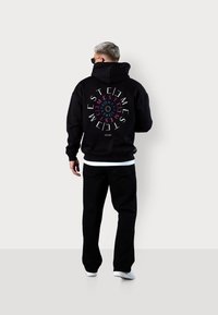 Mężczyzna o srebrnych włosach, ubrany w czarną bluzę z kapturem z okrągłym, kolorowym napisem, czarne spodnie, białe trampki i okulary przeciwsłoneczne, zwrócony plecami na jednolitym tle.