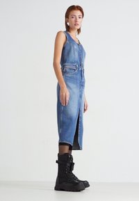 Robe en denim de couleur bleu clair, sans manches, avec des boutons sur le devant et une fente profonde. Associée à des bottes de combat noires. Texture lisse, silhouette ajustée.