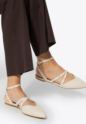 Nohy v béžových špičatých plochých sandálech s kotníkovými pásky a zlatými cvočky, v kombinaci s tmavě hnědými širokými kalhotami.