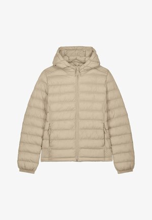 Vrouw staand, gekleed in een lichtbeige gewatteerd jack met capuchon, crèmekleurige cargobroek en witte plateau sneakers, tegen een effen achtergrond.