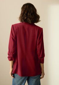 Sacou roșu cu design structurat, mâneci lungi suflecate la manșete și textură netedă, asortat cu blugi din denim.