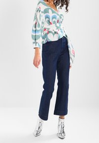 Nainen, joka on pukeutunut kuvioituun kietaisupaitaan, tummansinisiin leveisiin farkkuihin ja kiiltäviin hopeisiin nilkkabootseihin, seisoo valkoista taustaa vasten.