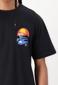 Sort bomulds t-shirt med en farverig grafik af et cocktailglas, sol, bjerge og bølger, trykt på venstre brystområde.
