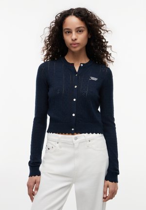 Vrouw met krullend haar die een marineblauwe cardigan met knopen en witte hoge taille broek draagt, staand tegen een witte achtergrond.