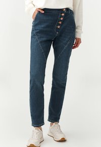 Dark blue denim jeans with a high waist, featuring a diagonal seam and five decorative buttons on the side. Smooth texture and slim fit.