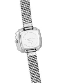 Montre en acier inoxydable argenté avec un boîtier carré à bords scallopés et un bracelet en maille. Elle présente des détails de marque et de mouvement gravés au dos.