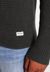 Dunkelgrauer Strickpullover mit einem strukturierten Muster. Hat lange Ärmel und ein Etikett an der Seite mit der Aufschrift "ONLY & SONS." Dazu hellblaue Jeans.