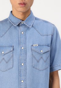 Šviesiai mėlynas trumpomis rankovėmis džinsinis marškiniai su dekoratyviniu siuvinėjimu, dviem priekinėmis kišenėmis su užsegimais ir Wrangler logotipo etikete.