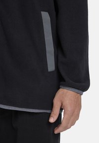 Pullover in pile nero con accenti grigi, dotato di tasca laterale, tessuto testurizzato e cuciture decorative attorno ai polsini e all'orlo.