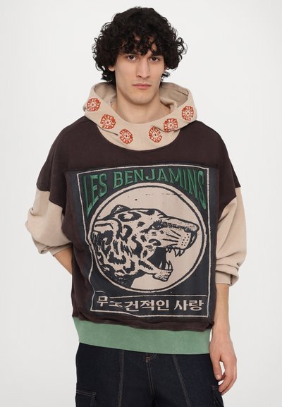 Haut à capuche de couleur marron foncé et beige avec un graphique de tigre, un texte vert et des accents décoratifs sur la capuche. Offre une coupe décontractée.