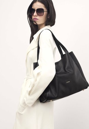 Borsa a mano in pelle nera con una texture morbida, caratterizzata da dettagli del logo oro e una forma strutturata. Il modello indossa un cappotto bianco e occhiali da sole grandi.