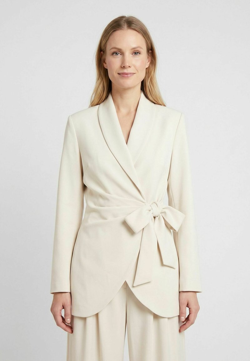 Femme portant un blazer portefeuille couleur crème avec des manches longues et un grand nœud noué à la taille, se tenant devant un fond uni.