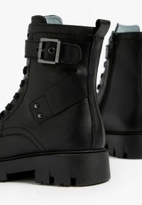 Schwarze Knöchelstiefel aus glattem Leder, mit einem Schnallenriemen, Schnürfront und dicker Gummisohle mit Profilmuster.