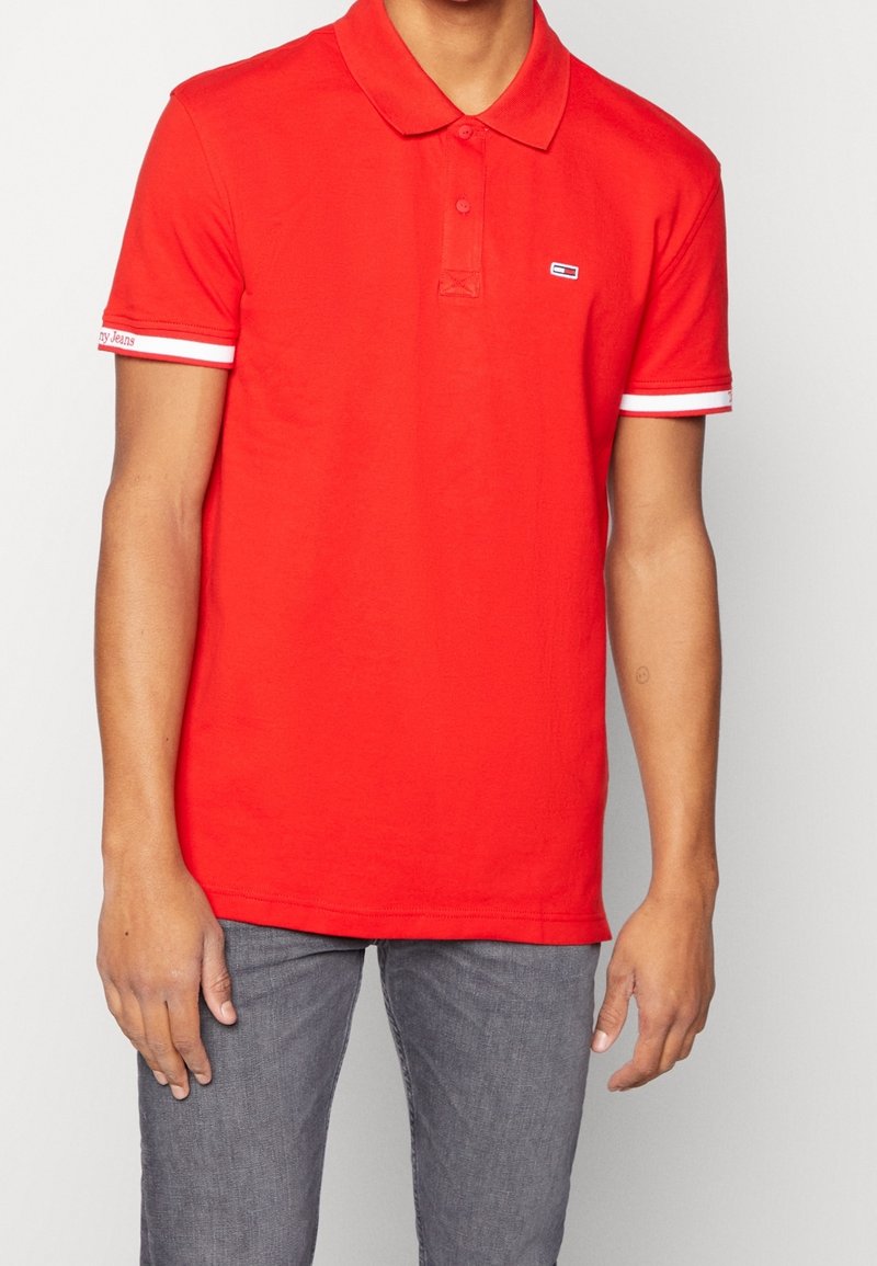 T-shirt polo rouge avec col, trois boutons et accents rayés blancs sur les manches. Présente un petit logo sur la poitrine. Fabriqué en coton.