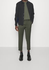 Mørk skjorte med knapper over en grøn rundhalset t-shirt, parret med grønne tapered bukser og sorte ankelstøvler i en minimalistisk setting.