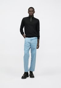 Mlad moški stoji, oblečen v črno pulover z zadrgo, svetlo modre kavbojke in črne velike čevlje, na enobarvni beli podlagi.