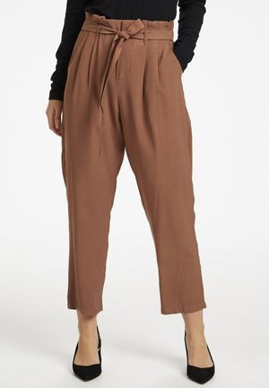 Frau trägt braune, hoch taillierte, schmal zulaufende Hose mit gebundenem Gürtel, schwarzes langärmeliges Oberteil und schwarze spitze Absatzschuhe, steht mit Händen in den Taschen.