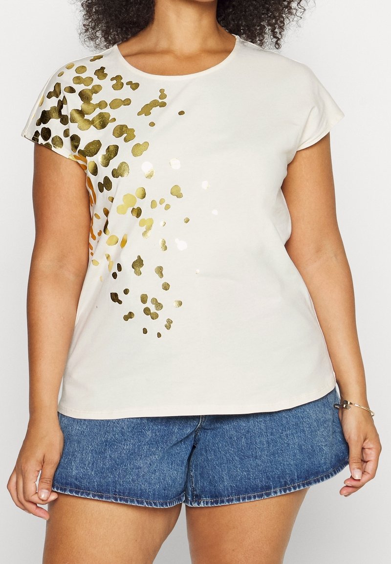 Kvinna iklädd en kortärmad beige topp med guldfolie-splatterdesign på vänster sida och blå denimshorts.