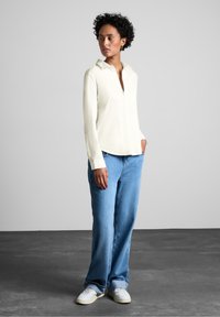 Lys beige langærmet skjorte med krave, parret med løse blå jeans og hvide sneakers på en grå baggrund.