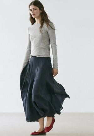 Donna con capelli lunghi indossa una maglia grigia a maniche lunghe, una gonna nera fluente e ballerine rosse, in piedi contro uno sfondo chiaro e semplice.