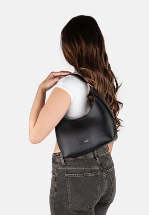 Γυναίκα με μακριά καστανά μαλλιά που φοράει άσπρο τοπ και μαύρα τζιν, κρατώντας μαύρη δερμάτινη τσάντα ώμου με χρυσό λογότυπο "RAID".