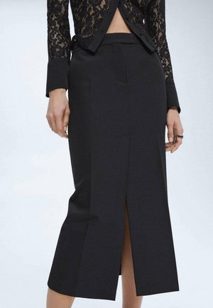 Jupe midi noire taille haute avec fente devant, portée avec un top en dentelle noire à manches longues, laissant apparaître partiellement le ventre et les mains avec des bagues.