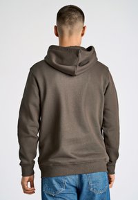 Brun hættetrøje med struktureret stof og ribbede manchetter, båret sammen med lyseblå denim jeans. Udsigten er fra bagenden.