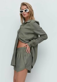 Woman wearing oversized olive green shirt and matching shorts, posing sideways with white rectangular sunglasses against a plain light background.