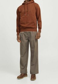 Sudadera marrón con el texto "ORIGINALS", con bolsillo canguro; combinada con pantalones grises de corte holgado y zapatos marrones con cordones.