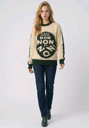 Joven mujer con cabello largo y rubio viste un suéter beige y verde oscuro con el texto "C'est non non non et non", vaqueros oscuros y botas verdes.