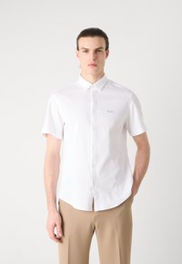 Camicia bianca a maniche corte realizzata in tessuto liscio, con colletto button-down e ricamo logo discreto, abbinata a pantaloni beige.