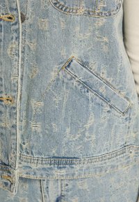 Jeansowa kurtka w jasnoniebieskim kolorze z postarzanym wykończeniem. Posiada dużą przednią kieszeń, metalowe elementy i widoczne detale przeszyć.