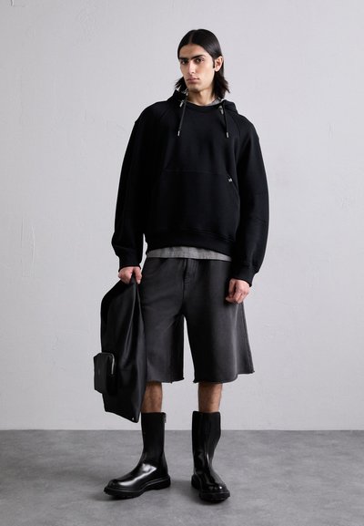 Črna oversize kapuca z vrvicami, siva plastična majica spodaj, temno sivi šorts, črni sijajni gležnarji in črna jakna v roki.