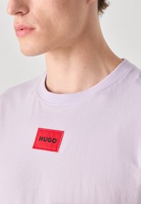 Levendula színű, pamut póló, kerek nyakkal, amelyen egy piros téglalap alakú folt található, rajta fekete hímzéssel a "HUGO" felirattal.