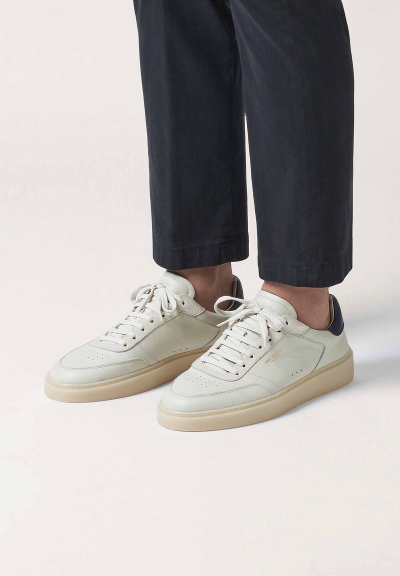 Zapatillas con un upper de cuero blanco, suela de goma marrón claro y detalle en el talón en azul marino. Presentan cordones redondos y detalles perforados en la puntera.