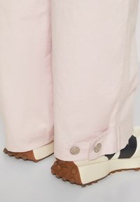 Pantalons en coton rose clair avec un ourlet retroussé, ornés de boutons en métal. Des baskets noires et blanches avec des semelles en caoutchouc brun texturé sont visibles.