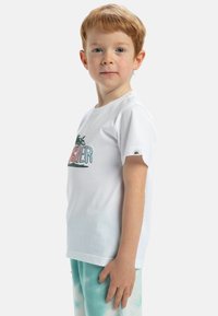 Camiseta blanca de algodón con un gráfico colorido de palmeras y texto, combinada con pantalones de tie-dye en verde menta. Mangas cortas y cuello redondo.