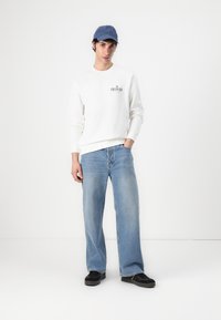 Hvit genser med logo, lyseblå vide jeans og svarte sko. Også med en blå caps og rett holdning mot en nøytral bakgrunn.