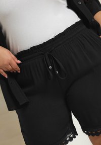 Mustat shortsit, joissa on joustava vyötärö, nyöri ja hopeiset helmet, teksturoitu kangas sekä pitsireunus helman yksityiskohtana.