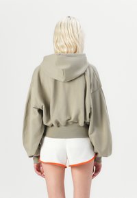 Sudadera grande de color verde oliva con capucha ajustable, dobladillo acanalado y mangas voluminosas, combinada con pantalones cortos blancos con ribete naranja.